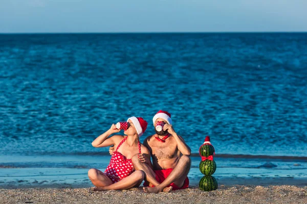 Bearded man and girl in Santa's clothes are drinking from cups on seashore, and snowman made of watermelons is standing nearby. Summer Christmas Celebration. — Stock Photo, Image
