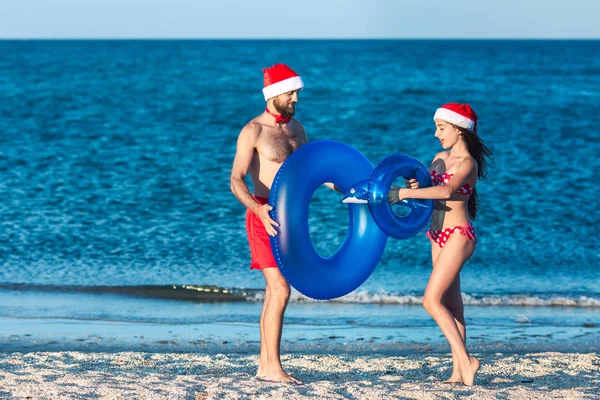 Brodaty facet i dziewczyna w kapelusze Mikołaja grać z nadmuchiwane kręgi i nadmuchiwane Delfin na plaży morskiej. — Zdjęcie stockowe