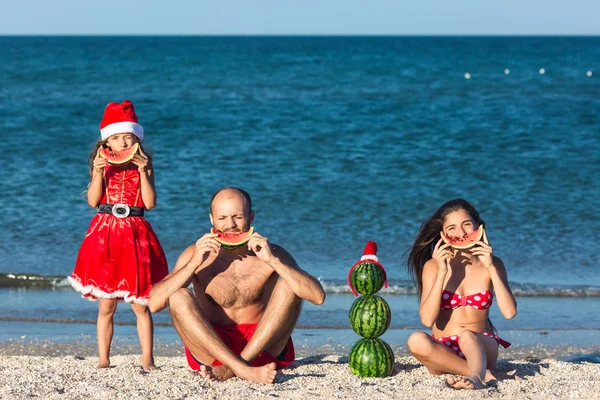 Merry family celebrates summer Christmas. father and two daughters eating watermelon on beach.