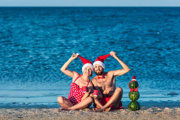 Brodaty mężczyzna i dziewczynka w Santa ubrania siedzą na plaży morskiej z kubków w rękach i bałwana z arbuz. Letnie święta Bożego Narodzenia. — Zdjęcie stockowe