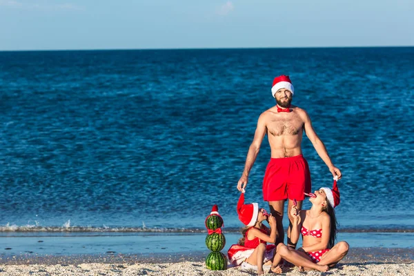 Young friendly fun family celebrates summer Christmas. father and daughters are sitting on beach with snowman from watermelons and holding cane lollipops.