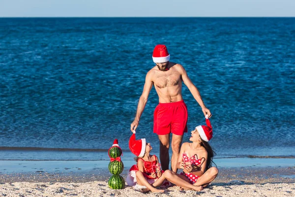 Young friendly fun family celebrates summer Christmas. father and daughters are sitting on beach with snowman from watermelons and holding cane lollipops. — Stock Photo, Image