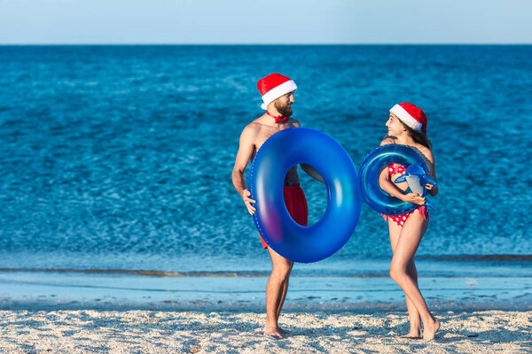 bearded guy and girl in Santa's hats play with inflatable circles and an inflatable dolphin on sea beach.