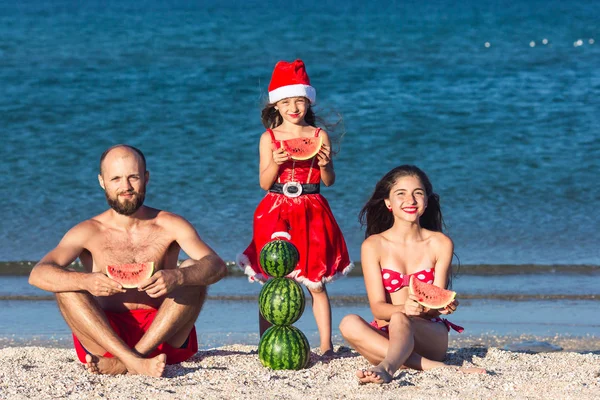 Wesoła rodzina świętuje letnie święta. Ojciec i dwie córki jedzą arbuza na plaży. — Zdjęcie stockowe