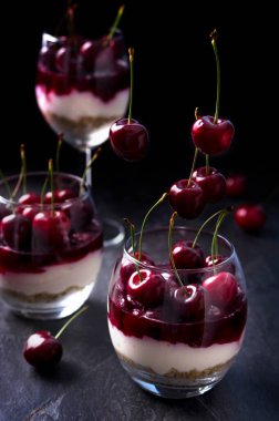 Bardakta kirazlı peynirli kek. Bisküvi, ricotta, krema, vişneli jöle ve taze meyveden yapılmış tatlı. Karanlık karamsar fotoğraf.