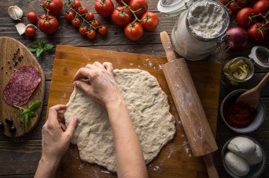 Bir kadın pizza hazırlıyor, hamuru yoğuruyor ve malzemeleri mutfak ahşap masasına koyuyor. Karanlık ve karamsar, havadan çekim.