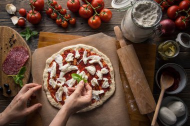 Bir kadın pizza hazırlıyor, hamuru yoğuruyor ve malzemeleri mutfak ahşap masasına koyuyor. Karanlık ve karamsar, havadan çekim.