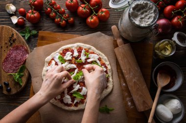 Bir kadın pizza hazırlıyor, hamuru yoğuruyor ve malzemeleri mutfak ahşap masasına koyuyor. Karanlık ve karamsar, havadan çekim.