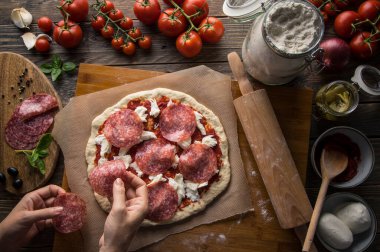 Bir kadın pizza hazırlıyor, hamuru yoğuruyor ve malzemeleri mutfak ahşap masasına koyuyor. Karanlık ve karamsar, havadan çekim.
