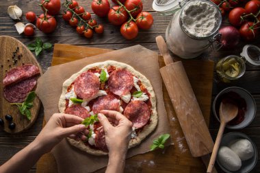 Bir kadın pizza hazırlıyor, hamuru yoğuruyor ve malzemeleri mutfak ahşap masasına koyuyor. Karanlık ve karamsar, havadan çekim.