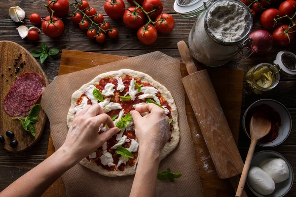 Bir kadın pizza hazırlıyor, hamuru yoğuruyor ve malzemeleri mutfak ahşap masasına koyuyor. Karanlık ve karamsar, havadan çekim.