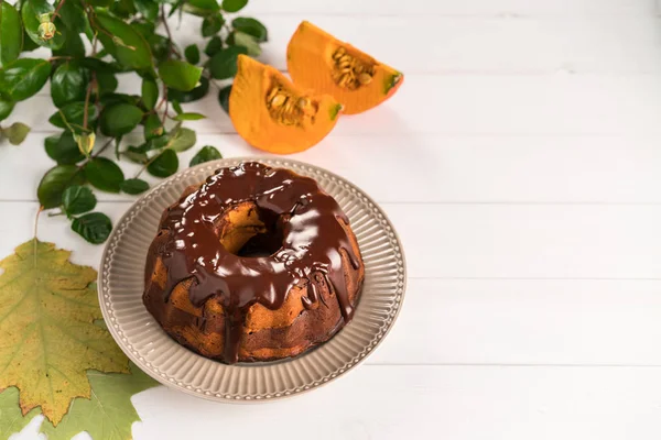 homemade chocolate and pumpkin cake, chocolate glaze on top, rustic plate, white wooden backdrop, decorated with leafs and pumpkin slices