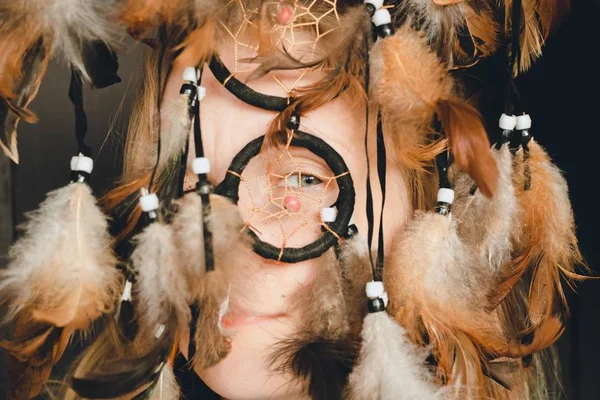 The girl looks through the ring of the dream catcher with one eye, feathers caress her tender face. With her hands she holds feathers that hang from the catcher dreams.