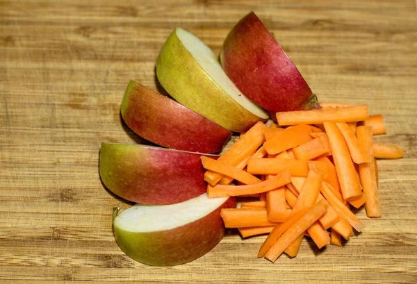 The apple of red and green hue is cut into pieces and laid out in a beautiful shape on a wooden surface. Next to the apple is sliced carrot orange shade.