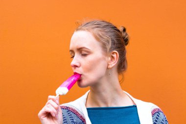 Güzel ve genç kız pembe bir renk dondurma yiyor ve sahiptir. Buz buzlanmanın tadını çıkarmaktan gözlerini kapadı. O turuncu bir gölge arka plan karşı duruyor.