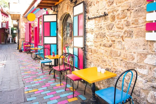 Bir şehir caddesinde bir kafenin aydınlık ve renkli iç. Dar sokak yolu çeşitli renklerle boyanmış taşlarla kaplıdır. Duvar boyunca masa ve parlak renklerde sandalyeler vardır.