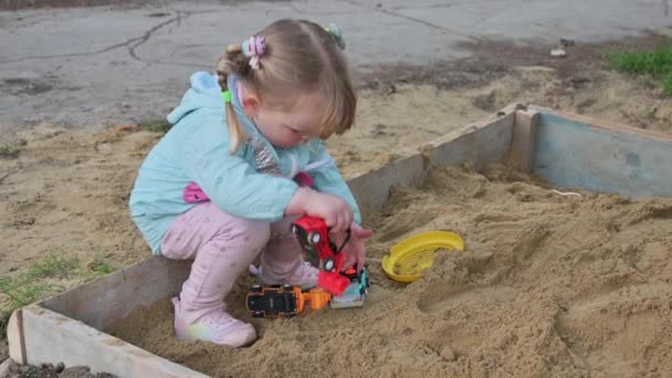 Petite fille jouant avec des voitures jouets dans un bac à sable — Video