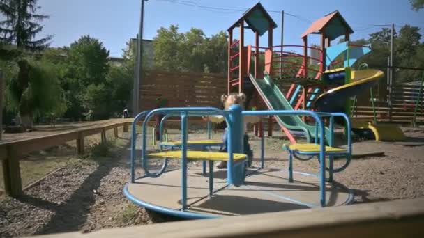 Little cute blond girl spinning on a merry go round at playground — Stock Video