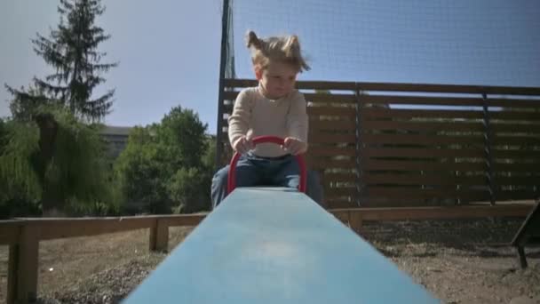 Liten vacker blond flicka rider teeter totter på lekplatsen — Stockvideo