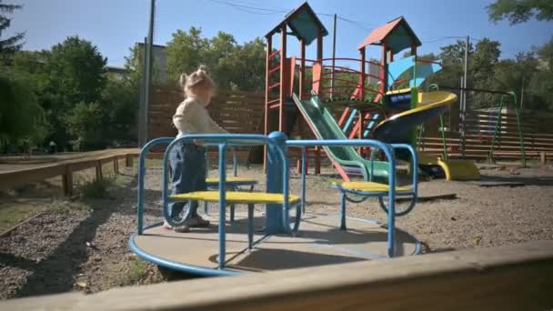 Kleines süßes blondes Mädchen dreht sich auf einem Karussell auf einem Spielplatz — Stockvideo