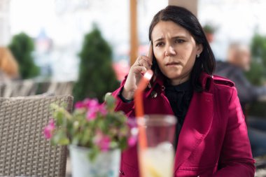 Açık hava kafeteryasında otururken cep telefonuyla konuşan pembe ceketli güzel bir kadının görüntüsü.