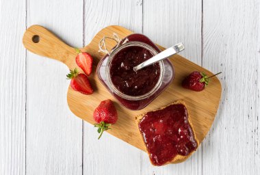 Cam kavanozda ev yapımı çilek reçeli ve beyaz ahşap arka planda bir kesme tahtasının üzerinde taze çilek ile bir tost ekmeğinin üzerine yayılmış. Kırsal biçim.