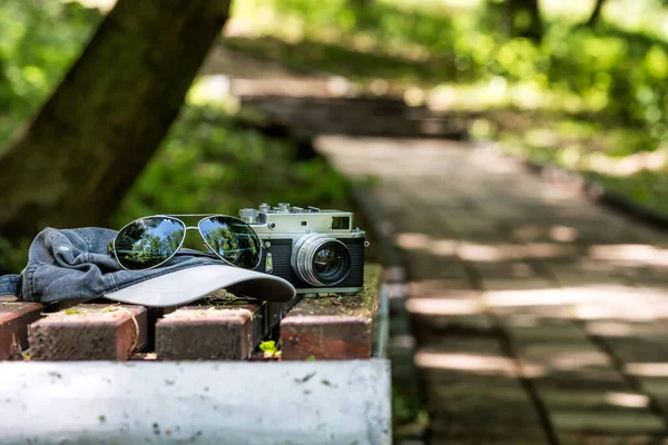 Parkın önündeki bankta eski fotoğrafçı aksesuarları. Rangefinder eski kamera, güneş gözlüğü ve beysbol şapkası.