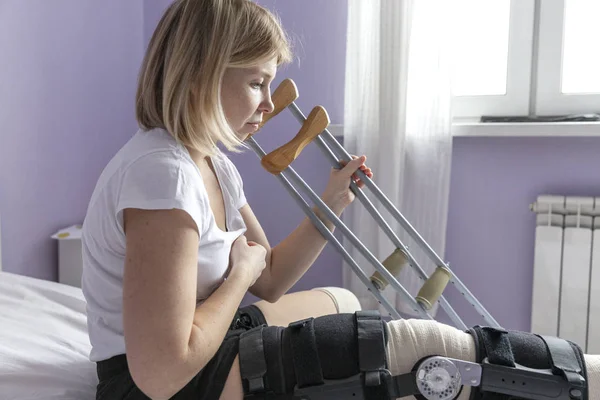 脚にオルソシスを持つ若い女性がベッドに座っています。松葉杖で立ち上がろうとする。術後期間. — ストック写真