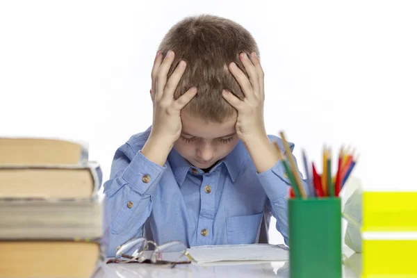 Söt ledsen skolpojke sitter vid bordet med böcker och anteckningsböcker. Isolerad på en vit bakgrund. — Stockfoto