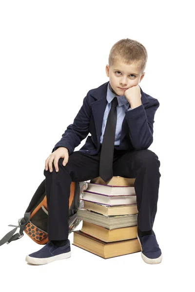 Söt ledsen skolpojke sitter på en bunt med böcker. Isolerad på en vit bakgrund. — Stockfoto