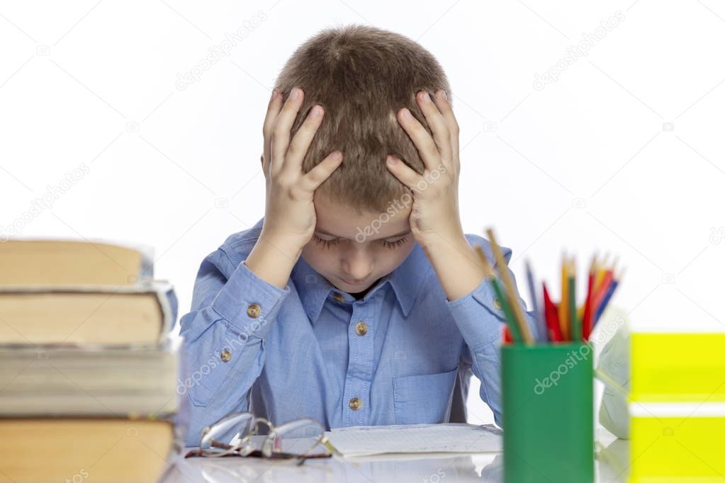 Cute sad schoolboy sitting at the table with books and notebooks. Isolated on a white background.