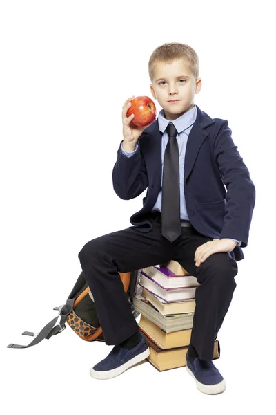 School jongen in een pak met een appel in zijn hand zittend op de boeken. Geïsoleerd op een witte achtergrond. — Stockfoto