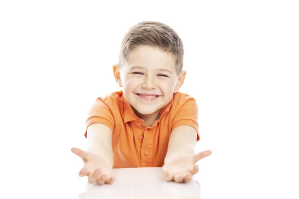 주황색 티셔츠를 입은 귀여운 웃음 학교 연령의 소년이 테이블에 앉아 팔을 뻗어 있습니다. 흰색 배경에 격리. — 스톡 사진