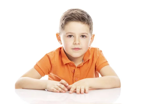 Netter Schuljunge in orangefarbenem T-Shirt, der am Tisch sitzt. isoliert auf weißem Hintergrund. — Stockfoto