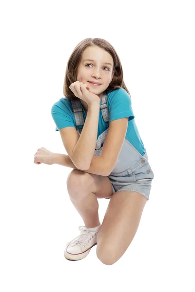 Smiling teen girl in denim overalls squats. Isolated on a white background. — Stock Photo, Image