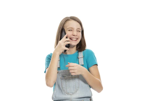 Cute teen girl in denim overalls talking on the phone and laughing. Isolated on a white background. — Stock Photo, Image