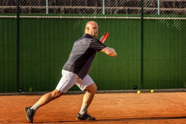 Orta yaşlı kel bir adam açık hava sahasında tenis oynar. Güneşli bir gün.