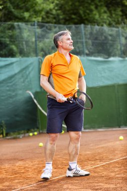 Orta yaşlı bir adam güneşli bir yaz gününde doğal toprak yüzeyi olan bir kortta tenis oynar.. 