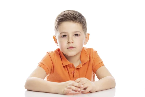 Am Tisch sitzt ein ernsthafter Junge im Schulalter in einem knallorangefarbenen Polo-T-Shirt. Nahaufnahme. isolirvoan auf weißem Hintergrund. — Stockfoto