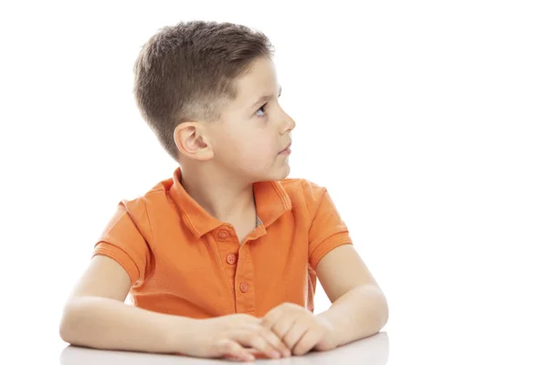 Poważny chłopiec w wieku szkolnym w jasnym pomarańczowym koszulce Polo siedzi przy stole i patrzy na bok. Zbliżenie. Isolirvoan na białym tle. — Zdjęcie stockowe