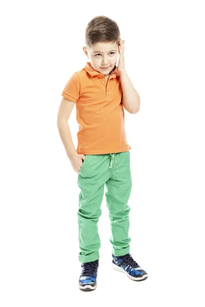 밝은 오렌지 색 폴로 티셔츠를 입은 귀여운 학령기 소년이 스마트폰으로 이야기하고 있습니다. 전체 높이. 흰색 배경에 격리. — 스톡 사진