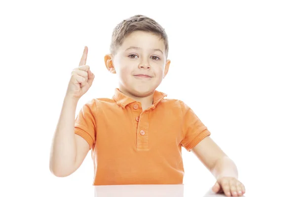 Een schattig school-jarige jongen in een helder oranje polo t-shirt zit aan tafel met zijn duim omhoog. Close-up. Isolirvoan op een witte achtergrond. — Stockfoto