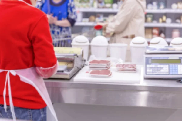 Um vendedor corta uma salsicha num supermercado. Borrão . — Fotografia de Stock