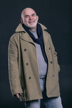 Gri sakallı yaşlı kel bir adam gülüyor. Şık kot pantolon ve bir ceket giymiş. Siyah arka plan. 