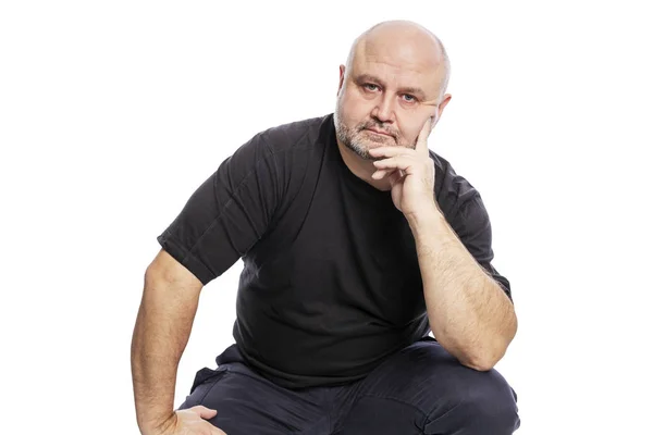 A serious bald middle-aged man in a black T-shirt is sitting with his hand in his face. Isolated over white background. — Stock Photo, Image