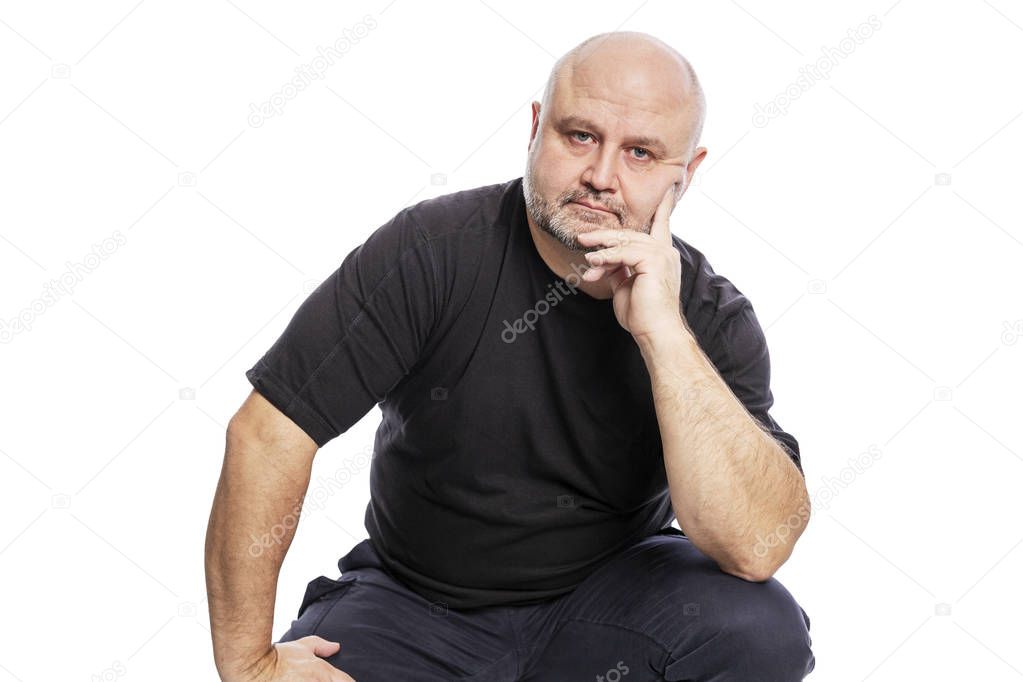 A serious bald middle-aged man in a black T-shirt is sitting with his hand in his face. Isolated over white background.