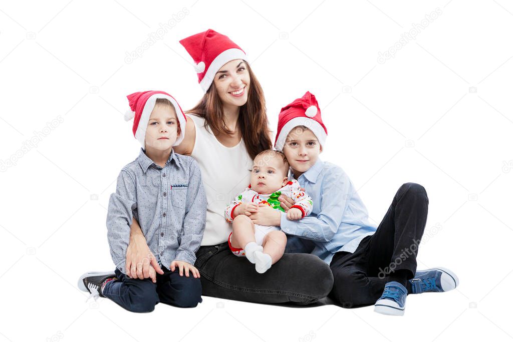 A smiling young mother with three young children in santa claus hats is sitting on the floor. Tenderness and family happiness. New Year and Christmas celebration. Isolated on a white background.