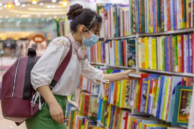 Tıp maskeli genç bir kız kitapçıdan bir kitap seçer. Bilgi ve eğitim. Koronavirüs salgını sırasında alınan önlemler..