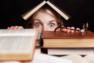 Genç bir kadın korku dolu bir kitap yığınından gözleri kocaman bir şekilde dışarı bakıyor. Eğitim ve eğitim. Siyah arka plan. Yakın plan..