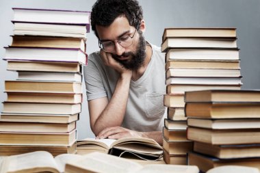 Gözlüklü ve sakallı genç bir adam büyük bir kitap yığınıyla bir masada oturuyor ve okuyor. Eğitim ve eğitim.